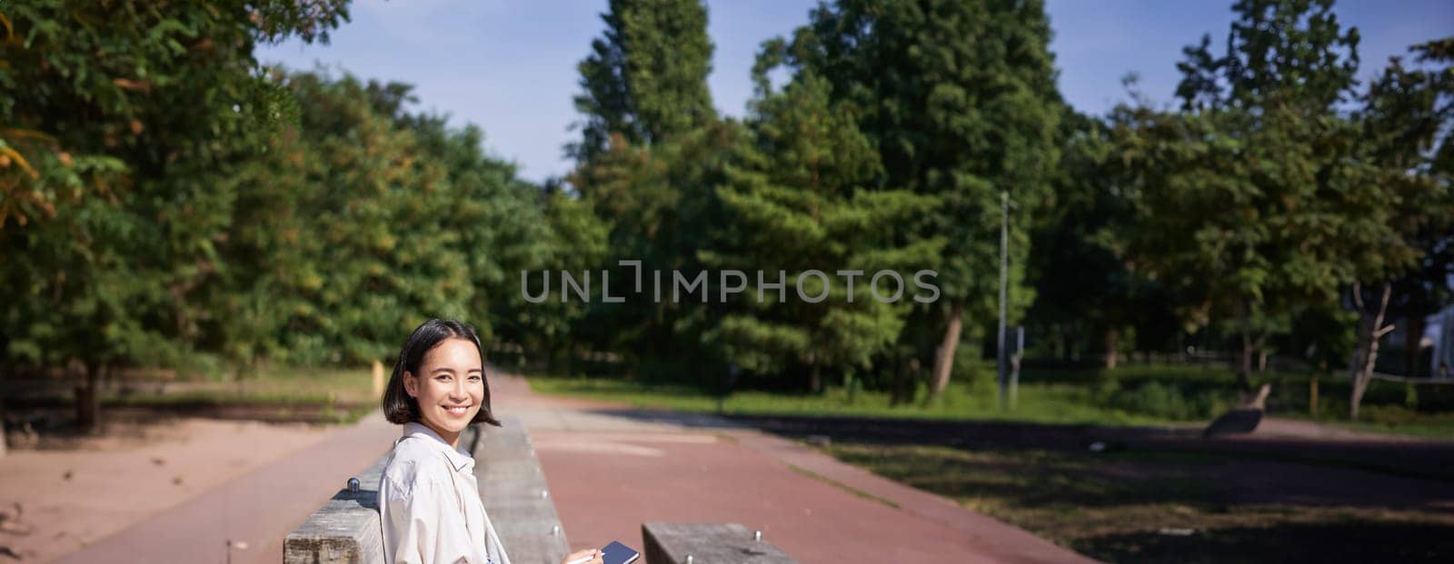 Asian girl, drawing in park, sitting on street on sunny day with digital graphic tablet and pen, smiling happily.
