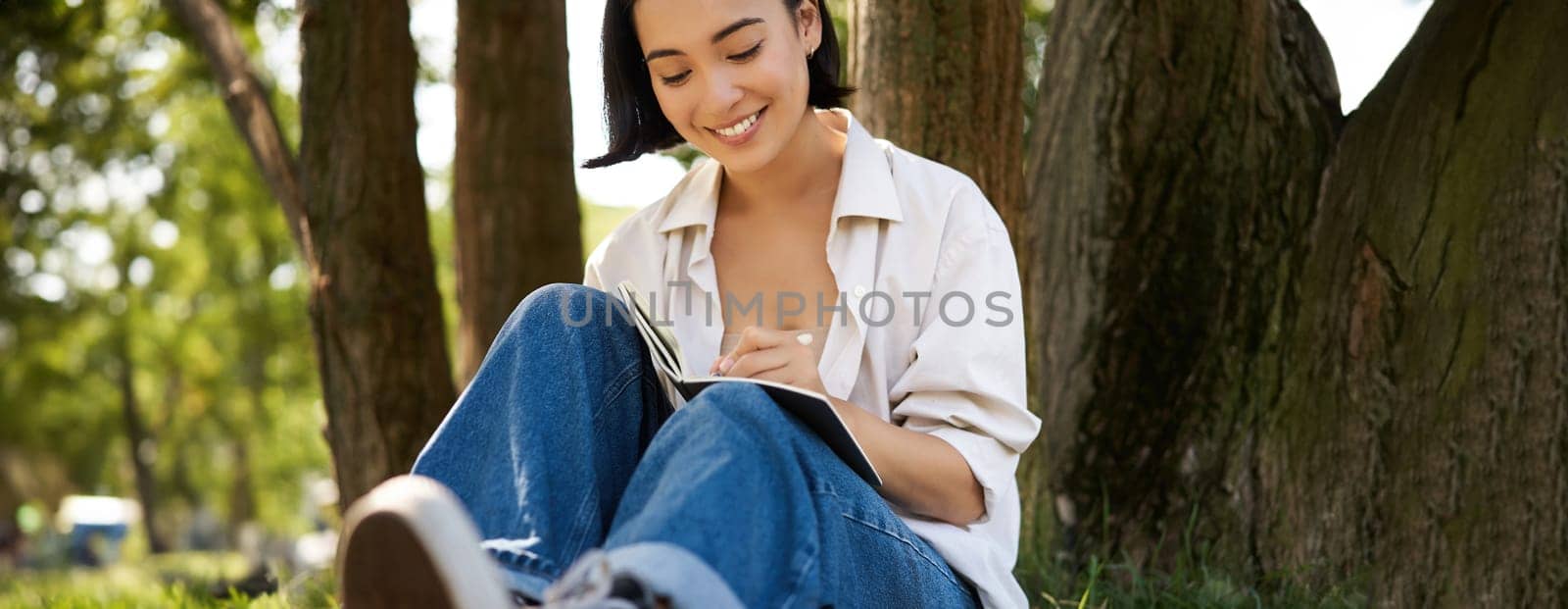 Portrait of happy asian girl sitting in park and writing in her diary. Young woman doing homework on fresh air, sitting near tree and smiling.
