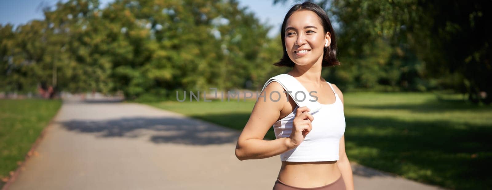 Smiling asian fitness girl holding towel on shoulder, workout in park, sweating after training exercises outdoors by Benzoix