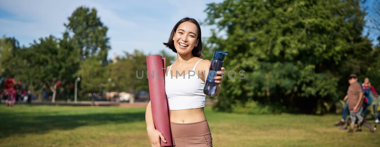 Portrait of happy asian girl, fitness woman giving you water bottle to drink after workout, standing with rubber mat for exercises in park.