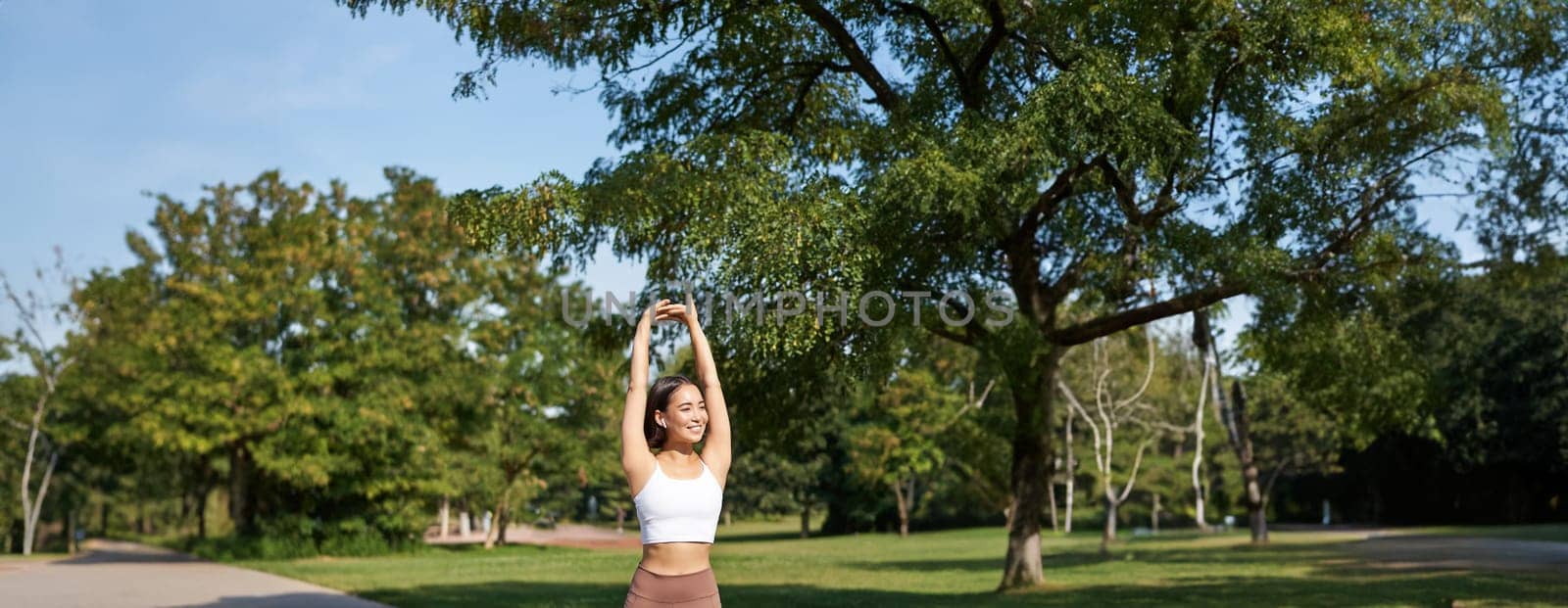 Smiling Asian girl stretching after good workout in park, listening music in wireless headphones, jogging outdoors by Benzoix