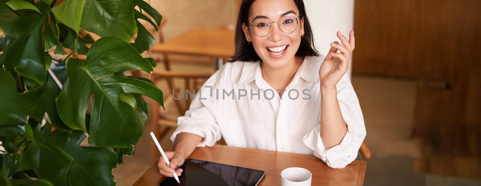 Young asian creative girl, sitting with digital tablet, drawing with graphic pen and smiling, doodling, drinking coffee in cafe.