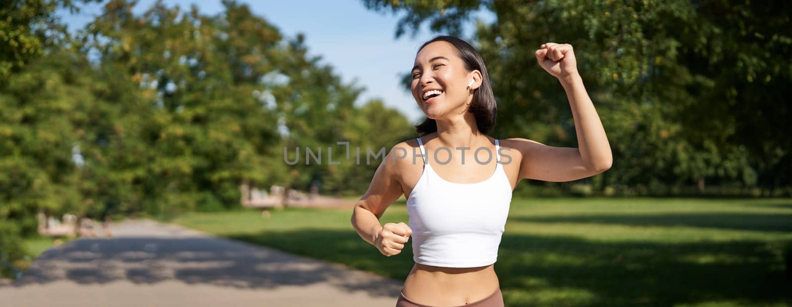 Happy fitness girl achieve goal, finish marathon, running with hands up, celebrating victory while jogging, triumphing in park by Benzoix