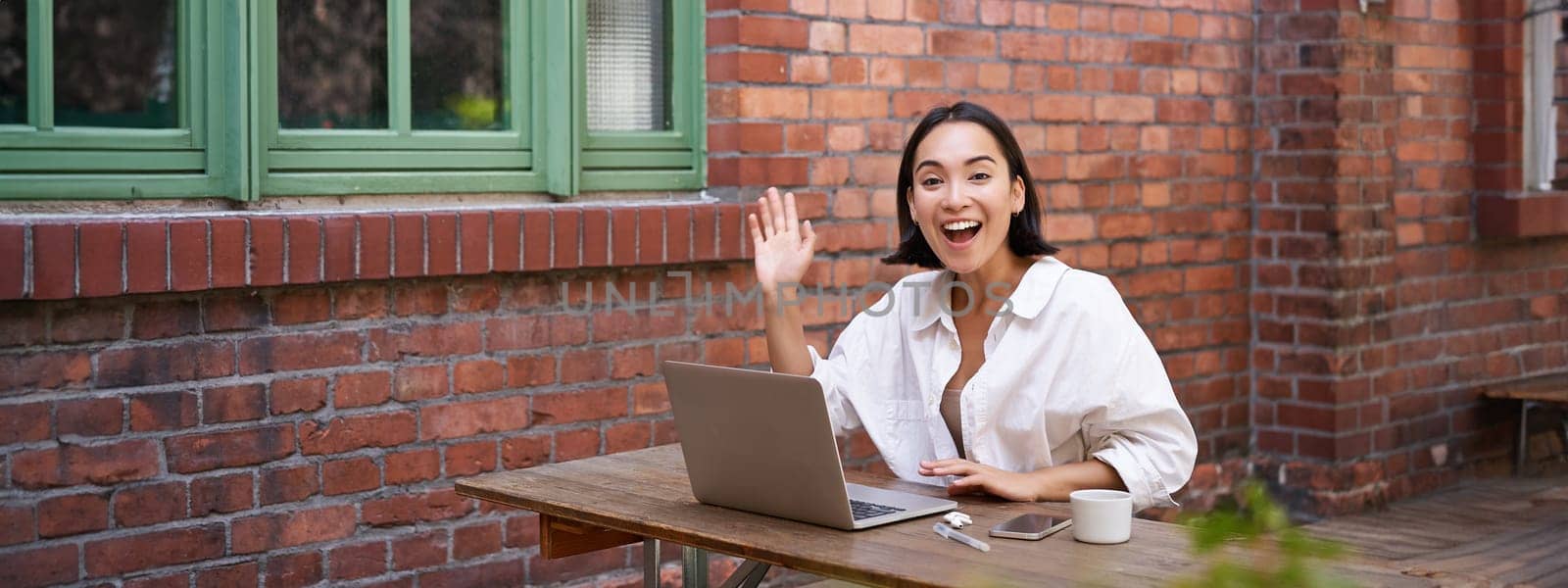 Friendly asian woman sitting with laptop, waving at you, saying hello, hi gesture, greeting you while working with computer.