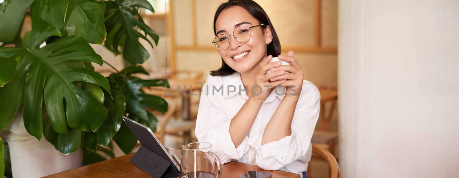 Smiling asian girl in glasses, woman working on remote, drinking coffee and using tablet. Copy space