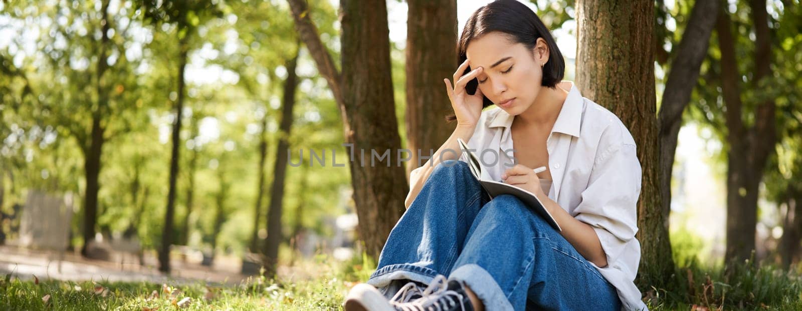Portrait of sad asian girl writing in her diary and feeling uneasy, sitting in park alone under tree, expressing her distress in notebook by Benzoix