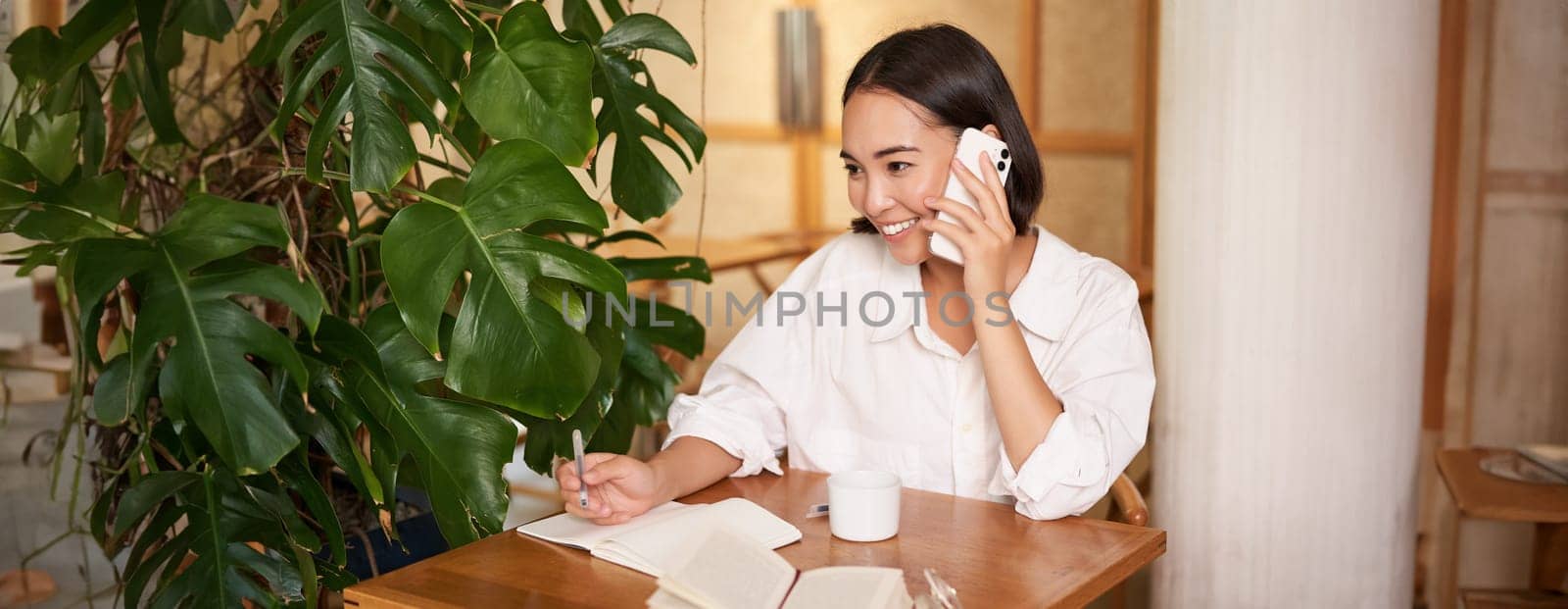 Working woman answer phone call in cafe, writing down, making notes while having conversation on telephone.