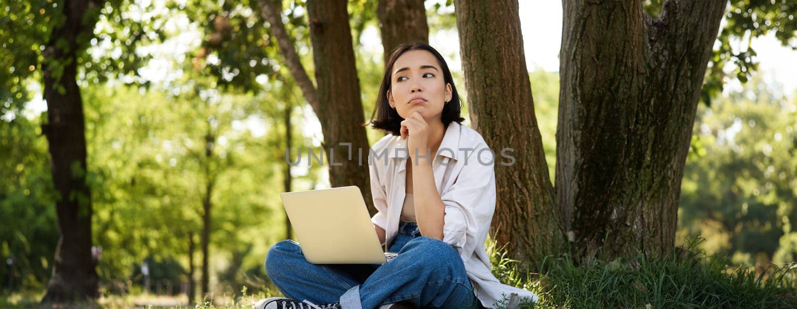 Portrait of young asian woman sitting in park near tree, working on laptop, using computer outdoors by Benzoix