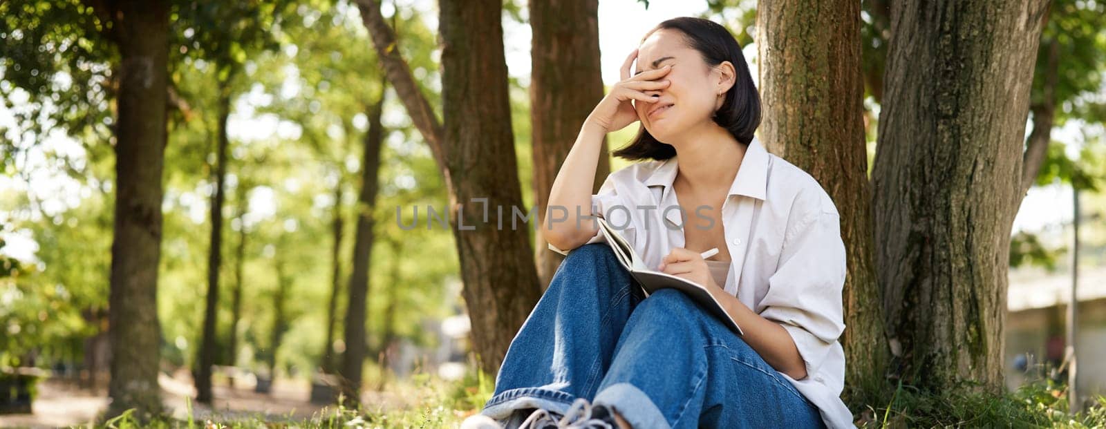 Sad girl crying while writing in her diary, sitting alone under tree in park with her notebook, sobbing and grimacing.