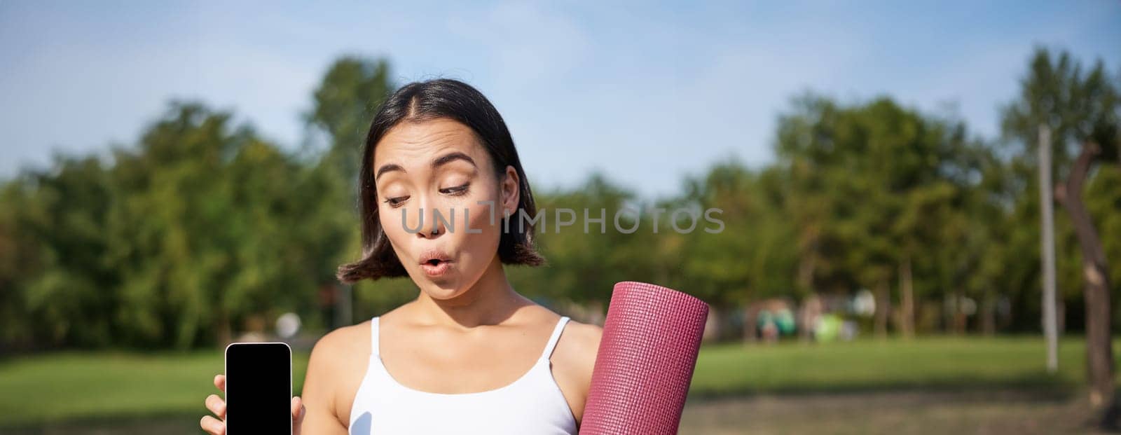 Excited fitness girl recommends application for sport and workout, shows phone screen, standing with rubber yoga mat in park after training session.