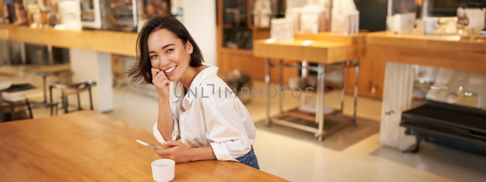 Portrait of young asian woman, sitting in cafe, holding smartphone, chatting and messaging while drinking coffee.