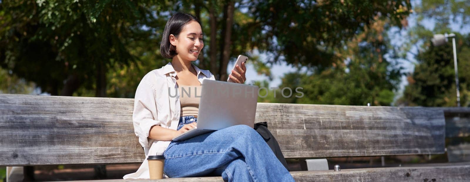 Excited young asian girl, looking at her smartphone, while sitting with laptop outdoors in sunny park.