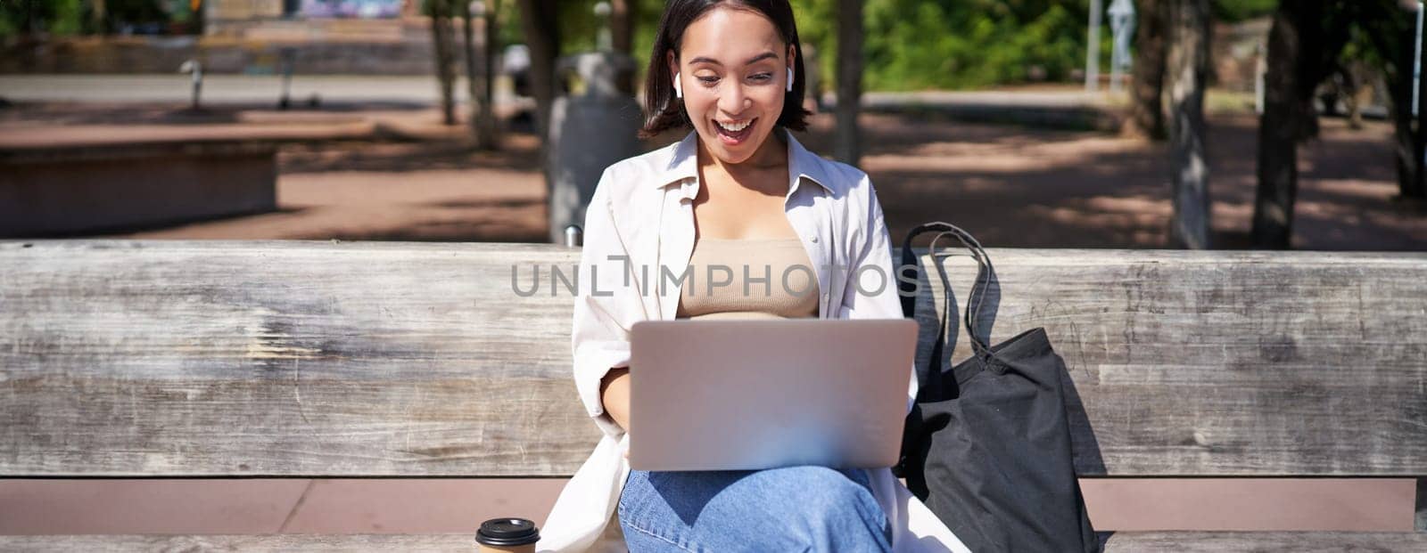 Happy asian girl chatting, having video call on laptop, sitting on bench in park and communicating, talking to someone online by Benzoix