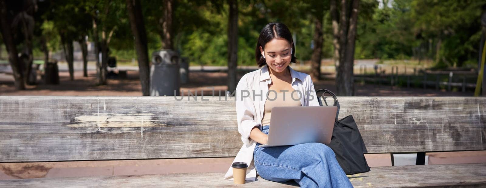 Portrait of korean woman sitting with laptop in park on bench, working outdoors, student doing homework, drinking takeaway coffee.