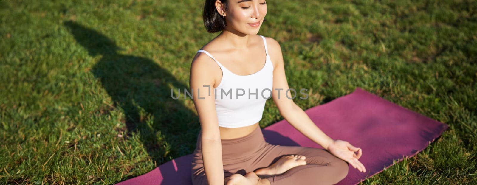 Portrait of smiling asian woman meditating, doing yoga on fresh air, relaxing on rubber mat, exercising in park, breathing air, being calm.