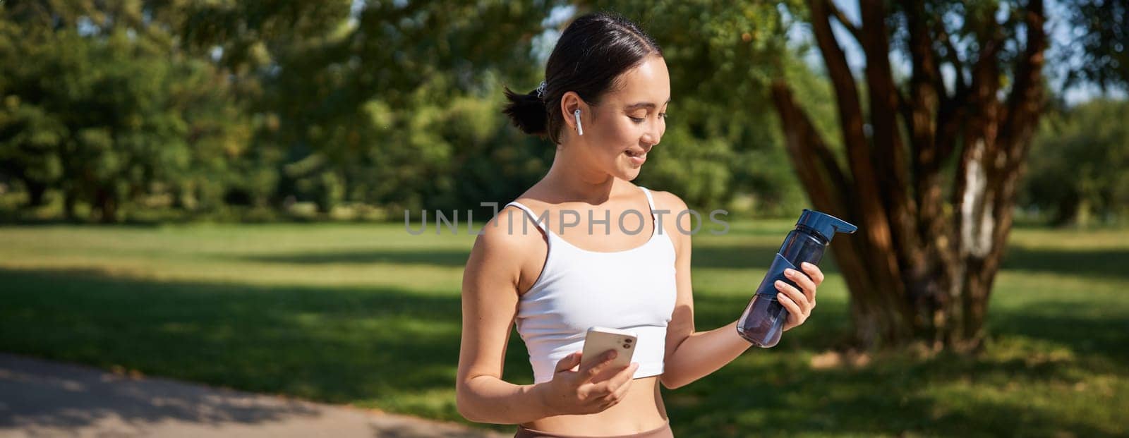 Portrait of fitness girl, runner drinking water, looking at smartphone, standing in park in middle of workout, sport training session.