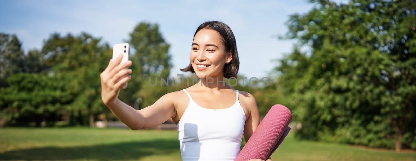 Sporty fitness girl takes selfie with rubber yoga mat in park, does workout and shares photos on social media.