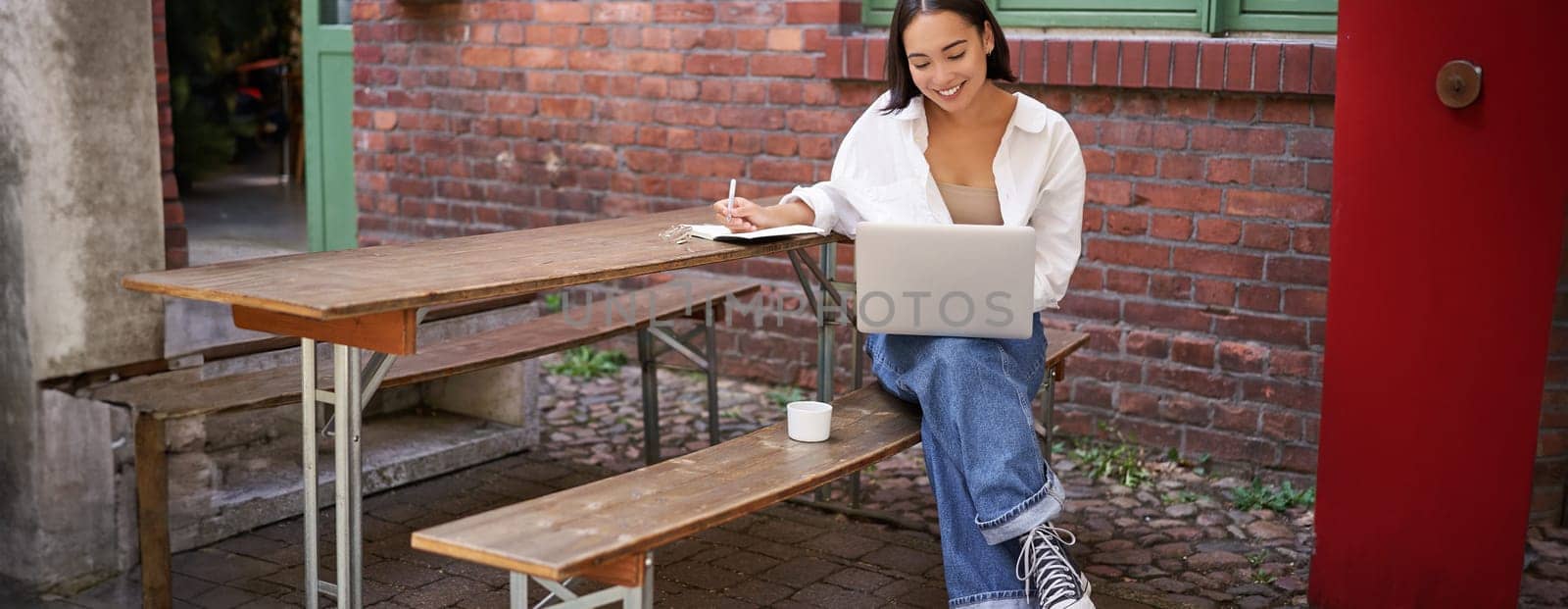 Happy asian student, girl writing down and studying smth on laptop, sits in outdoor cafe with coffee, working.