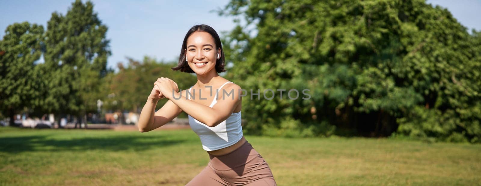 Wellbeing and sport complex. Young asian woman stretching, doing squats and workout on fresh air, smiling pleased.