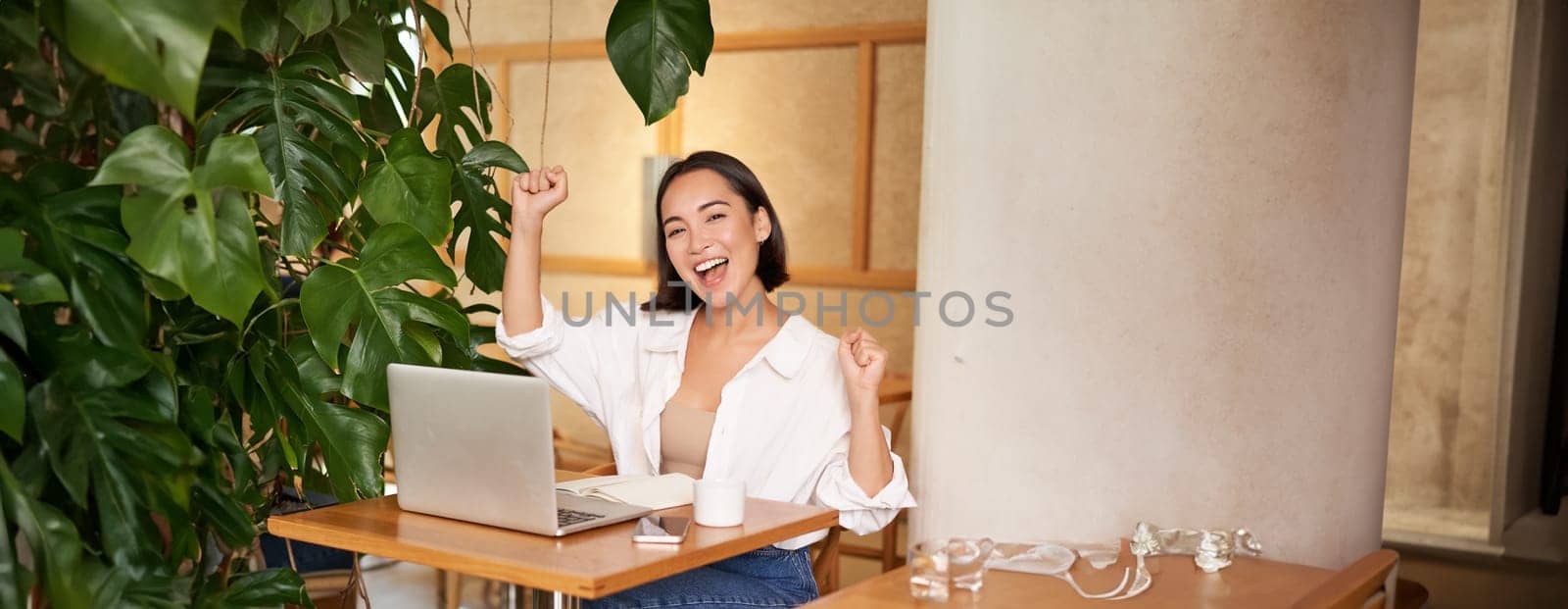 Happy young asian woman winning on laptop, receive good news, achieve goal at work, triumphing and smiling pleased, sitting in cafe.