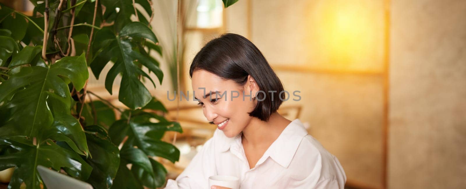 Student sitting in cafe with cup of coffee. Young asian woman working on laptop in restaurant, sitting with computer and smartphone.