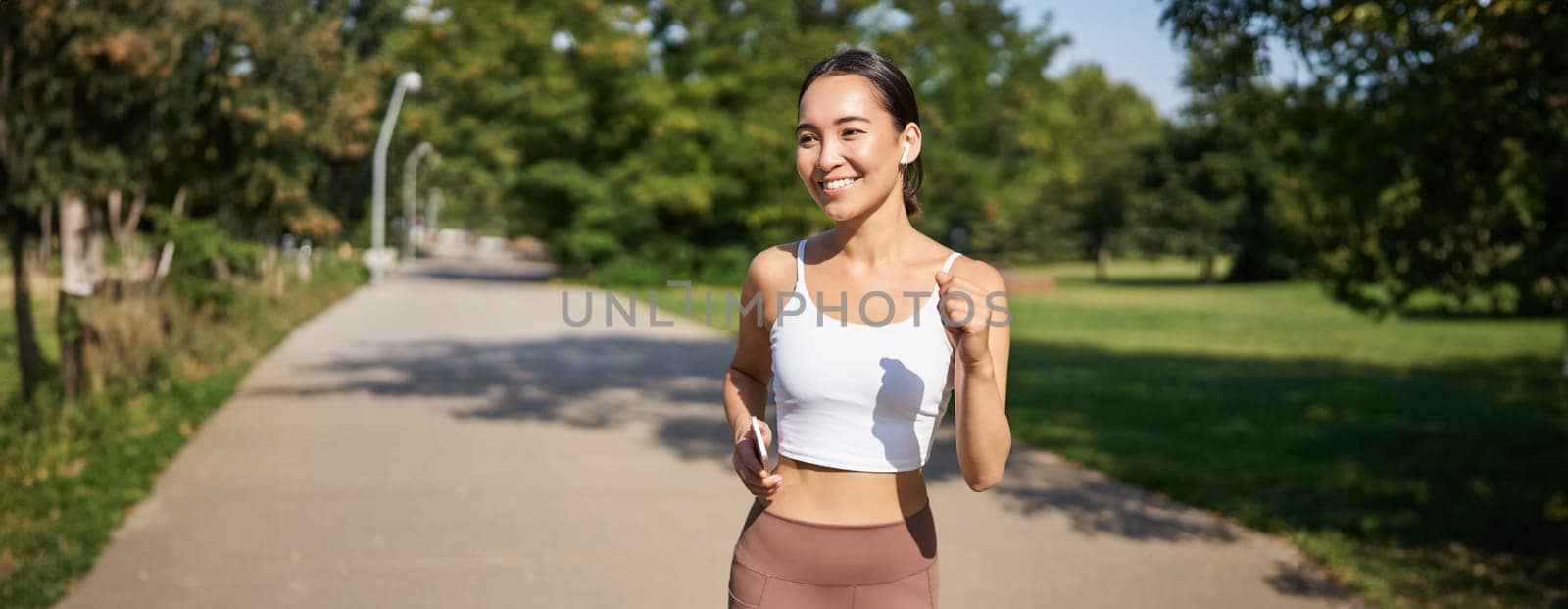 Happy smiling asian woman jogging in park. Healthy young female runner doing workout outdoors, running on streets.