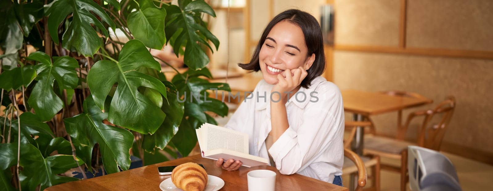 Romantic asian woman sitting with book in cafe, eating croissant and drinking coffee, reading and smiling, enjoying alone time by Benzoix