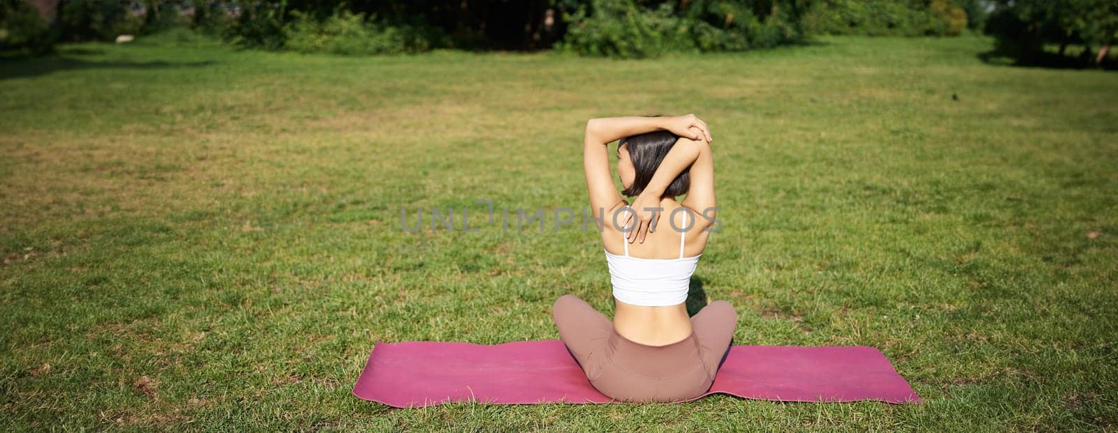 Rear view of sportswoman in middle of workout, stretching her amrs behind back, sitting on rubber yoga mat on lawn in park.