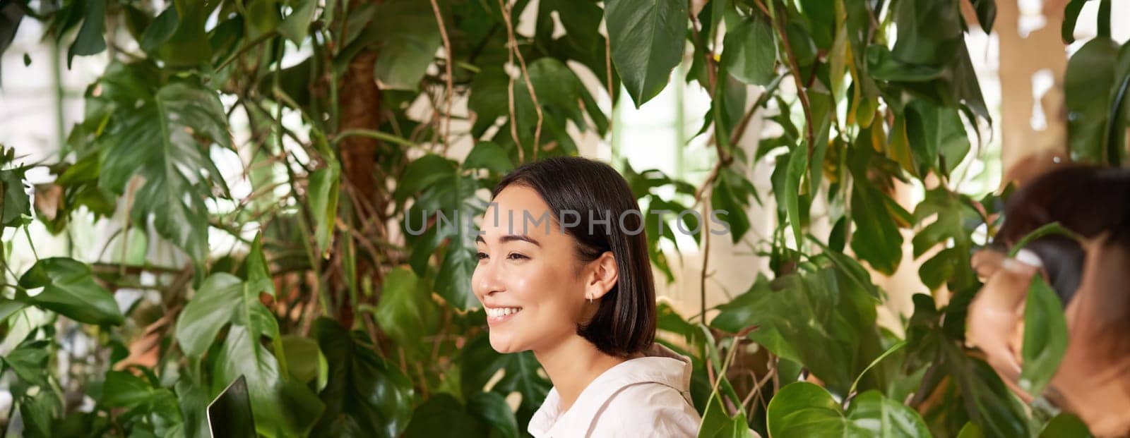 Vertical shot of beautiful woman sits in cafe with laptop, works or studies online at co-working space, smiling relaxed.