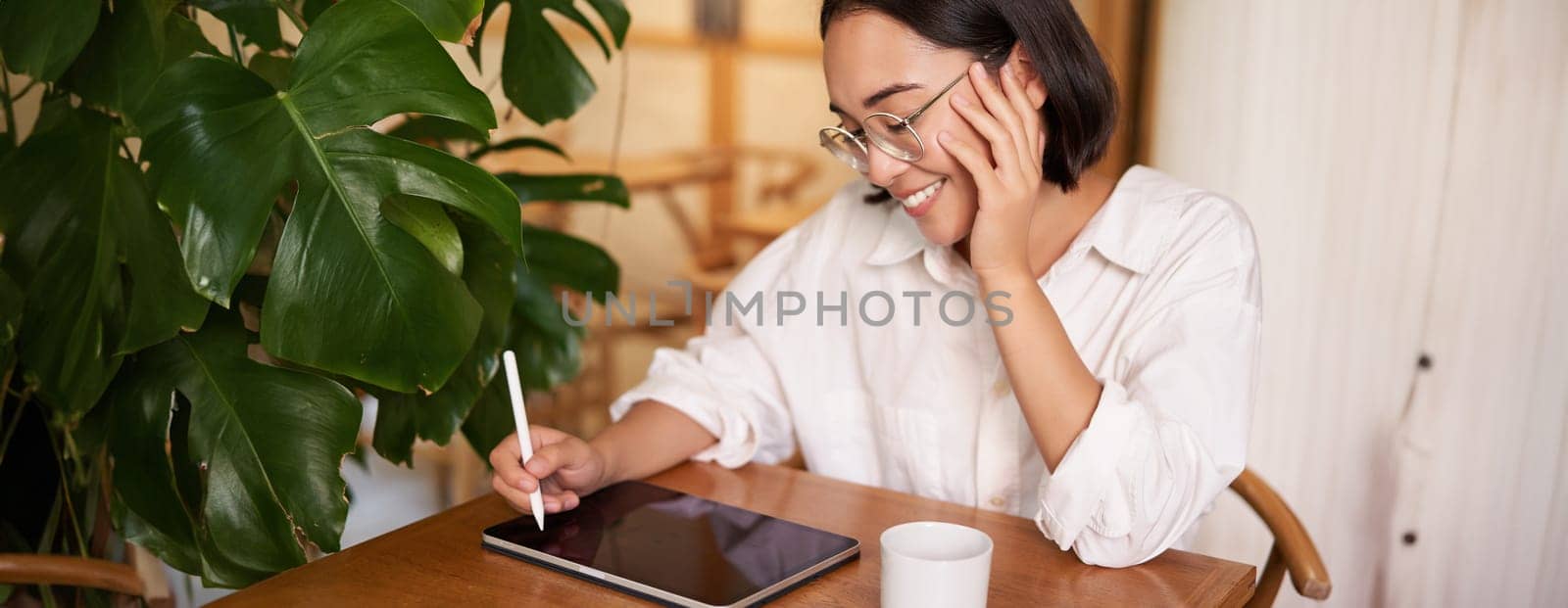 Young asian creative girl, sitting with digital tablet, drawing with graphic pen and smiling, doodling, drinking coffee in cafe.