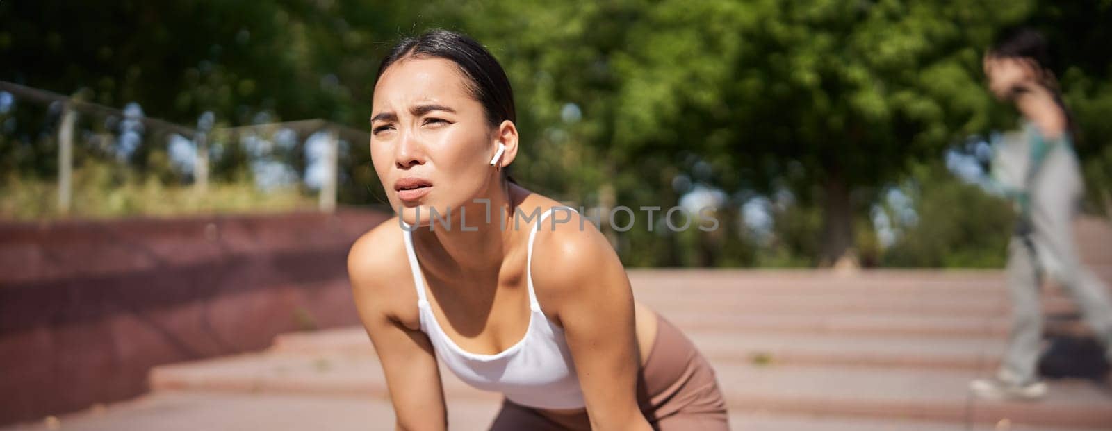 Tired young female runner, asian girl taking break during workout, stop jogging, panting while breathing, running in park.