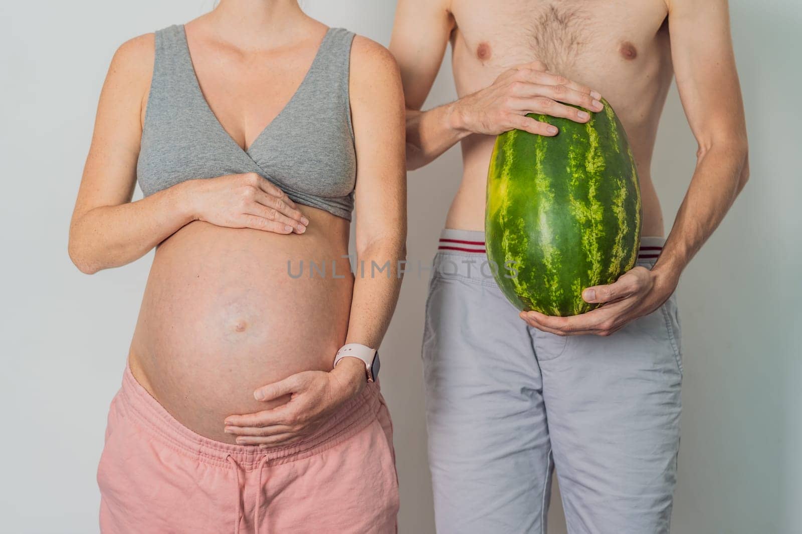 A humorous image: a pregnant woman and her husband playfully use a watermelon in place of a belly, comically highlighting the challenges of navigating with a pregnant bump by galitskaya