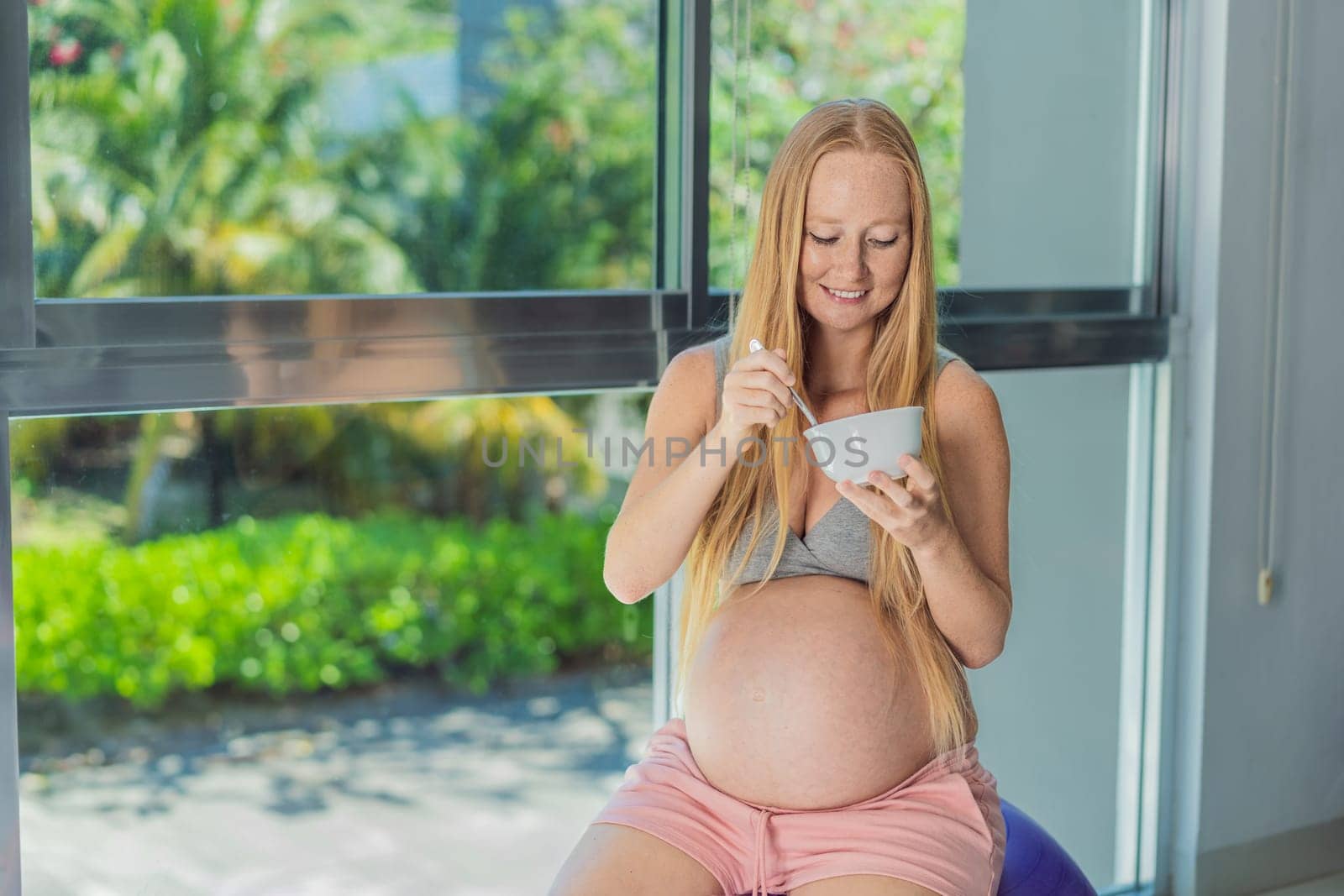 An expecting mother gracefully has breakfast on a fitness ball, illustrating the comfort it provides during pregnancy when traditional chairs may be less accommodating by galitskaya