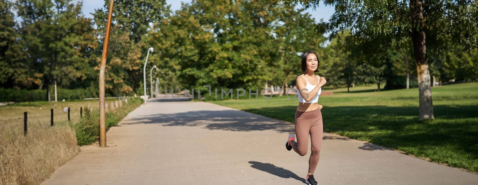 Young asian fitness woman running in park, jogging outdoors, smiling pleased.