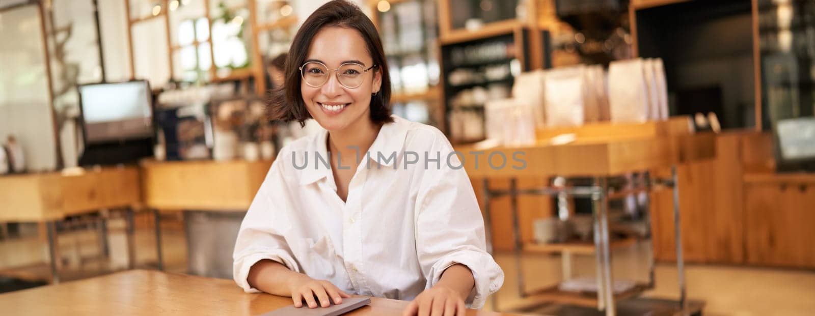 Young stylish asian woman, business owner in glasses, sitting in cafe with notebook, smiling at camera. People and lifestyle concept