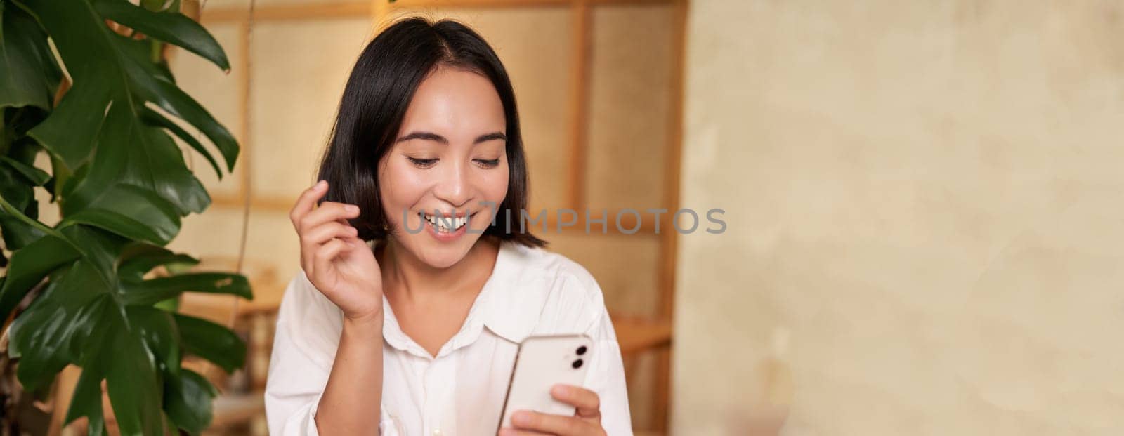 Portrait of modern girl sits with coffee in cafe, smiling while looking at smartphone, reading book in restaurant and using mobile phone app.