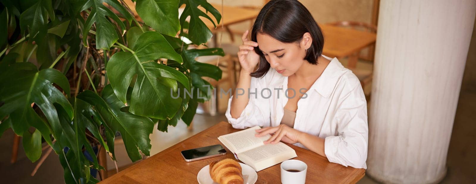 Beautiful young asian woman sitting in cafe with a book, eating croissant and reading, drinking cup of coffee. Wellbeing and self-care