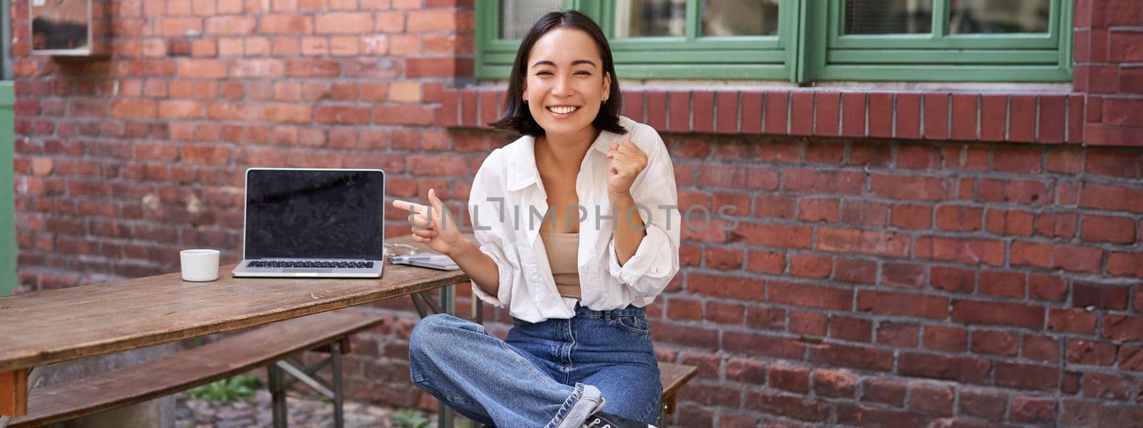 Happy korean girl with laptop, sitting in cafe, laughing and pointing at copy space on brick wall, showing advertisement.