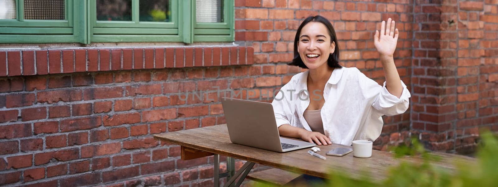 Friendly asian woman sitting with laptop, waving at you, saying hello, hi gesture, greeting you while working with computer.