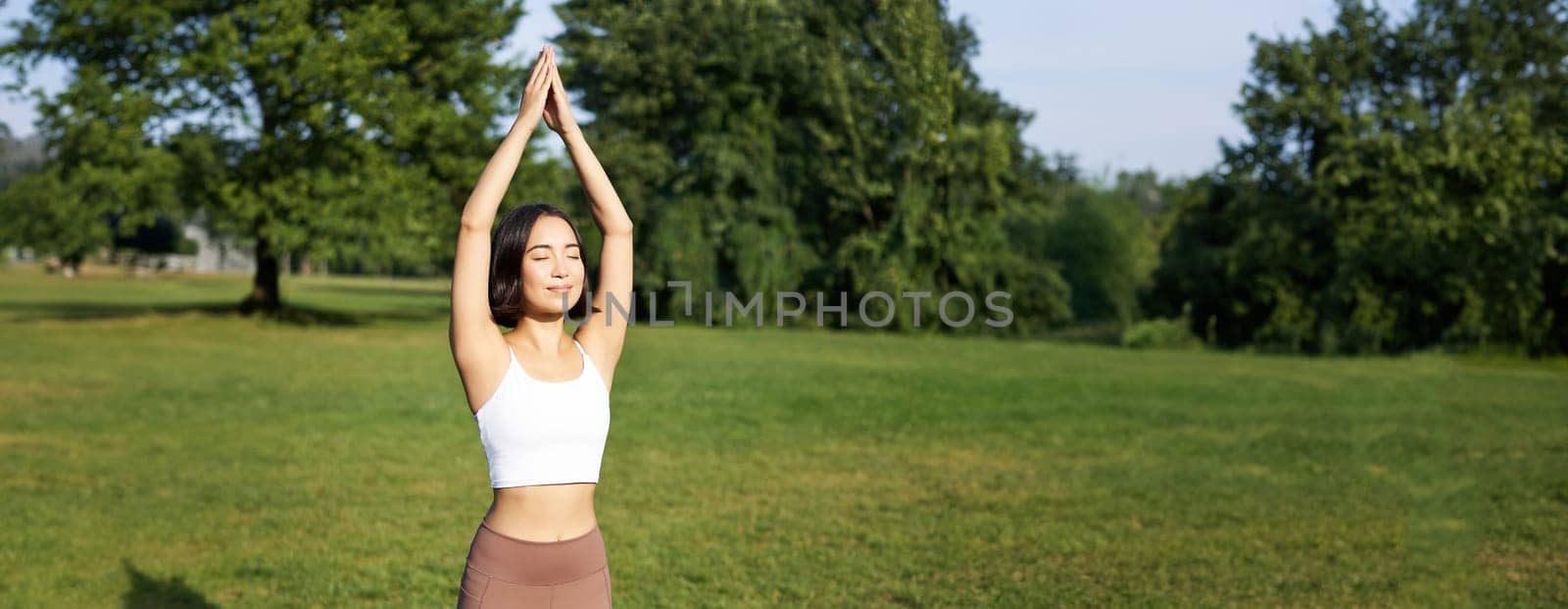 Smiling young fitness girl on rubber mat, workout in park, raising hands up in tree pose, doing yoga training on fresh green lawn.
