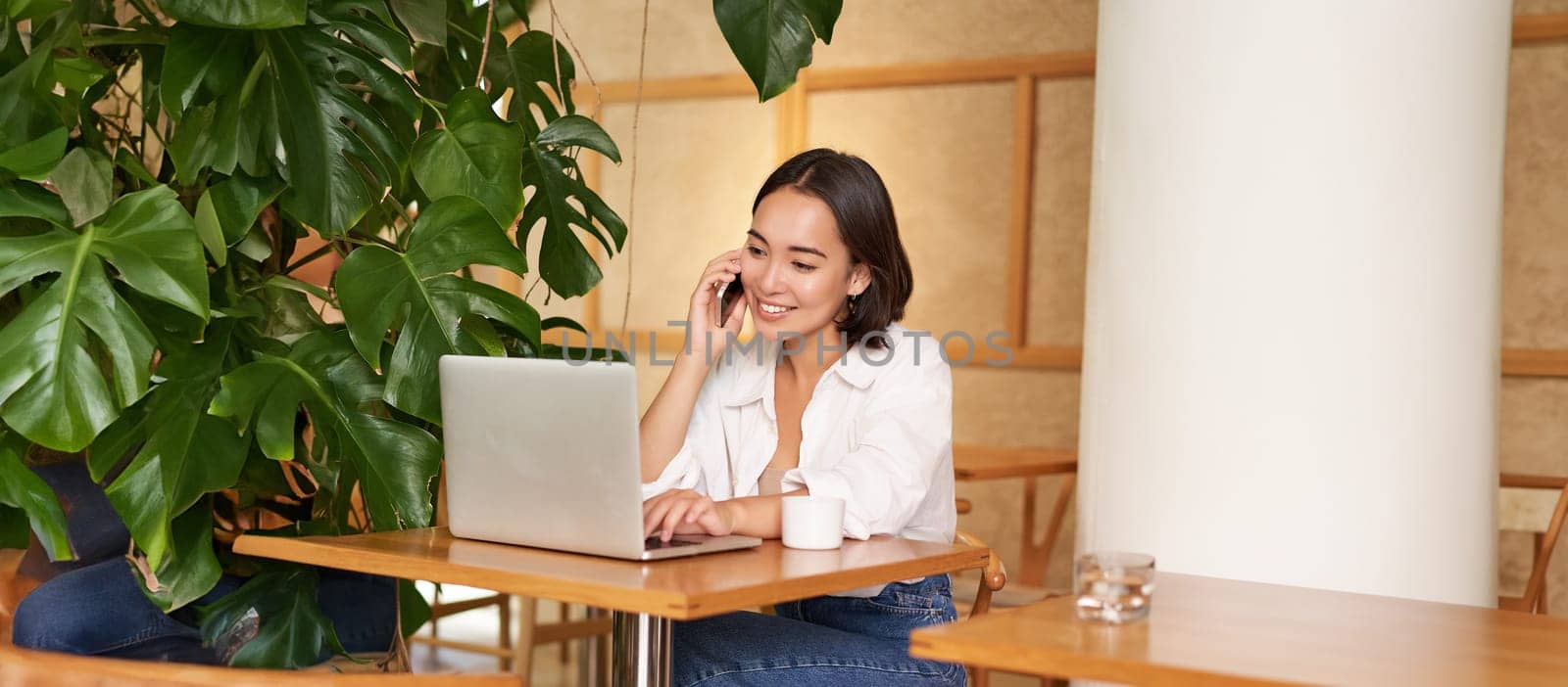 Stylish young woman answer phone call, sitting in cafe with laptop, working and taling on smartphone.