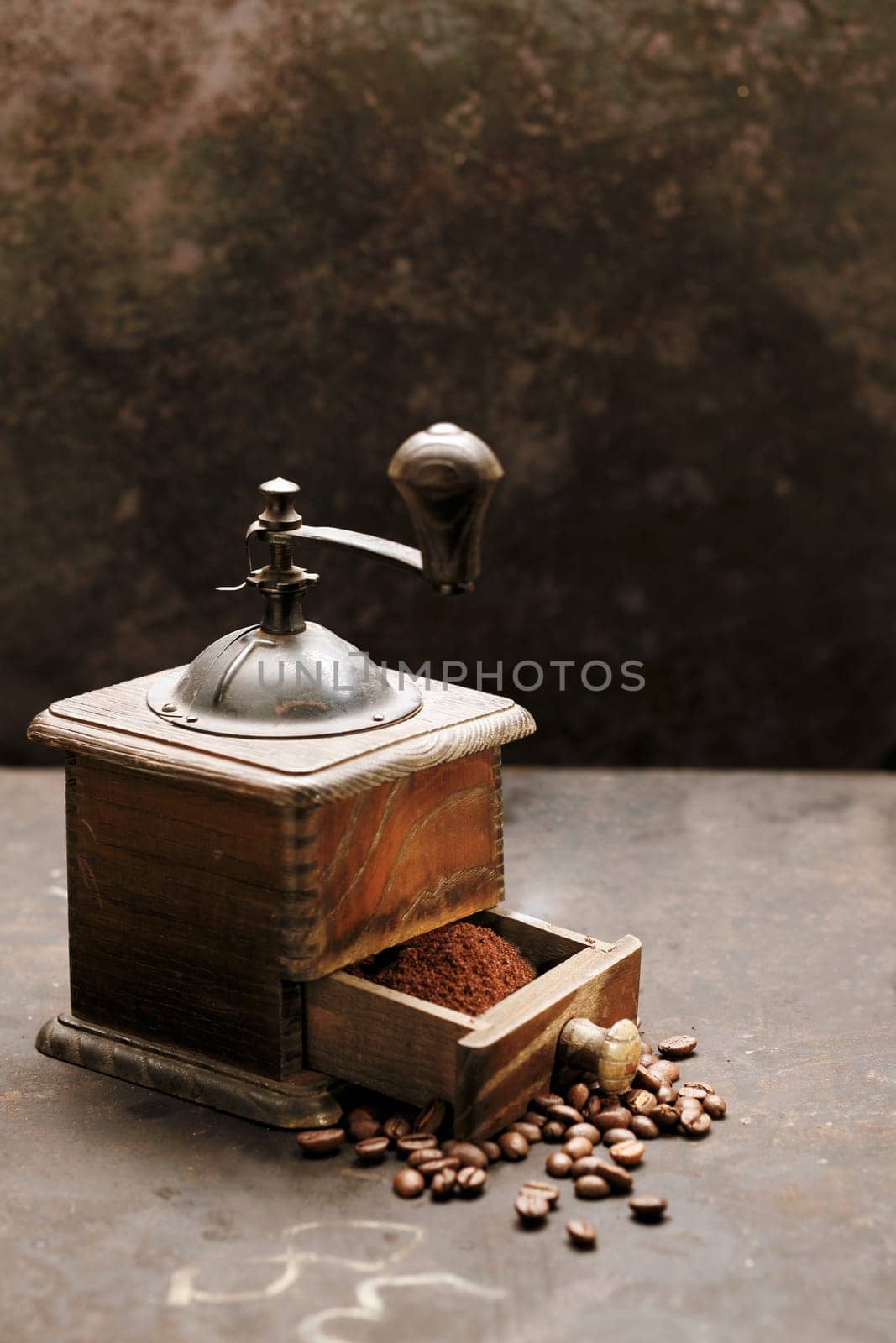 Old rustic wooden coffee grinder on a slate surface with scattered roasted coffee beans and freshly ground powder in the drawer ready to be used for filter coffee, espresso or cappuccino