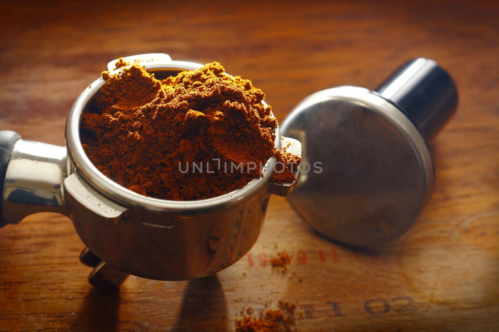 Freshly ground coffee overflowing over the rim of a metal filter onto a wooden surface with measuring spoon