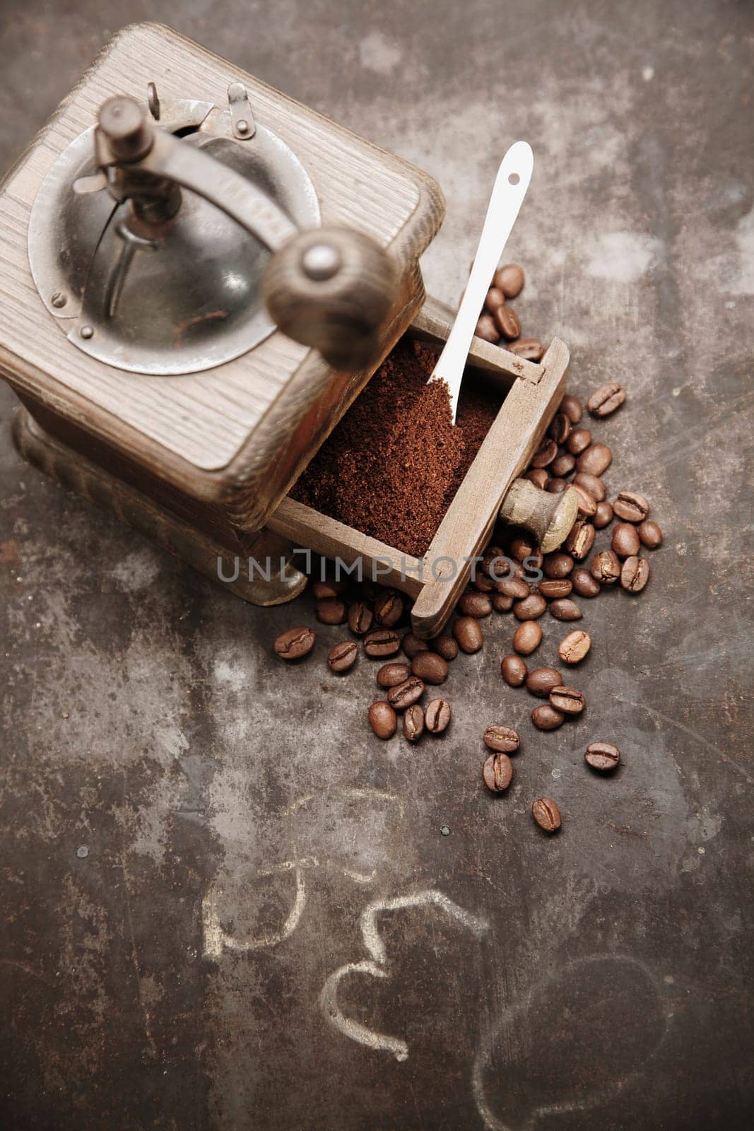 Overhead view of an old manual wooden coffee grinder with an open drawer filled with freshly ground coffee surrounded by scattered coffee beans on a grungy chalkboard with copyspace