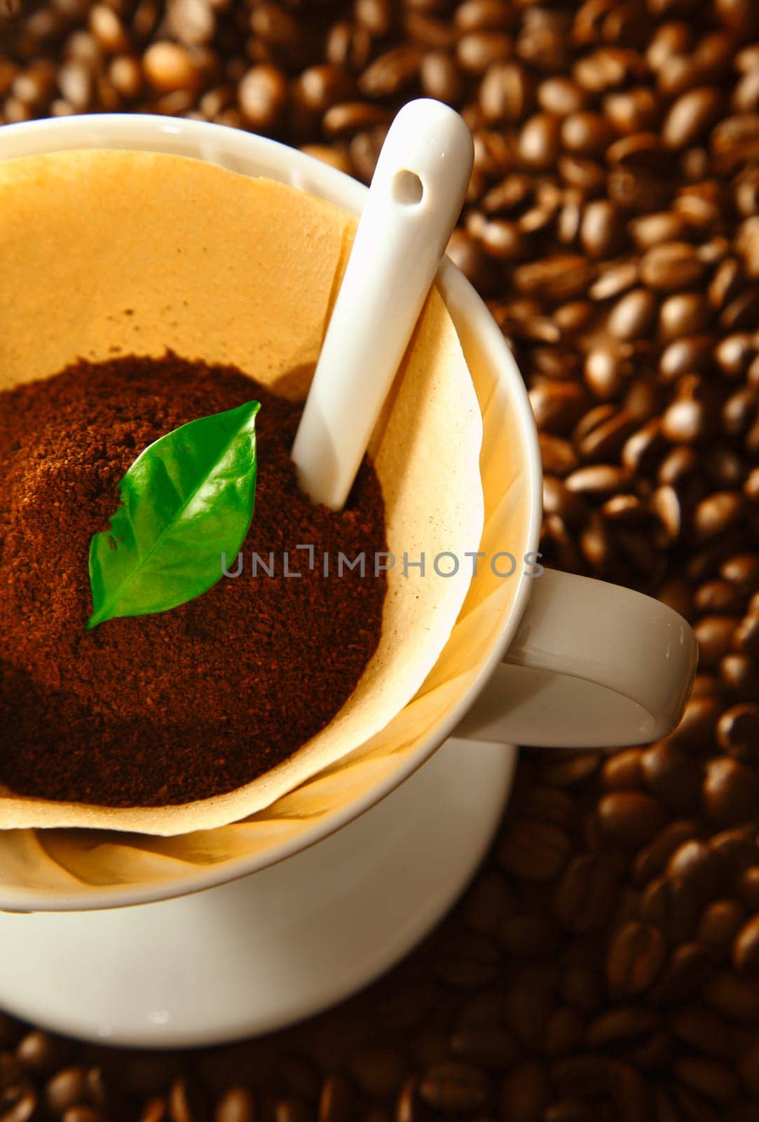 Fresh ground coffee and a green leaf in a portable filter funnel on a bed of whole roasted beans during preparation of a cup of filter codffee