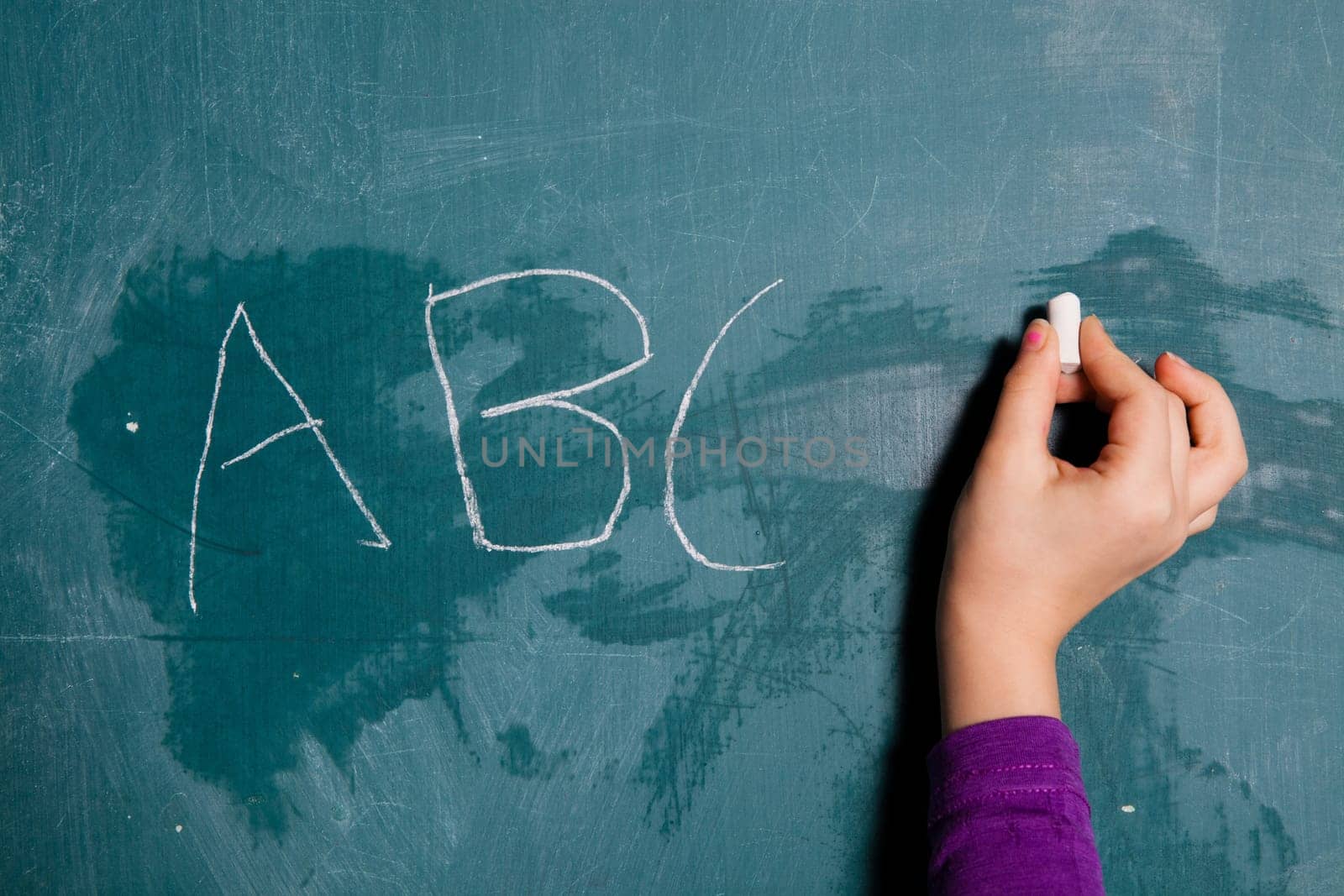 Hand of young child holding stick of white chalk to write letters of alphabet on chalkboard