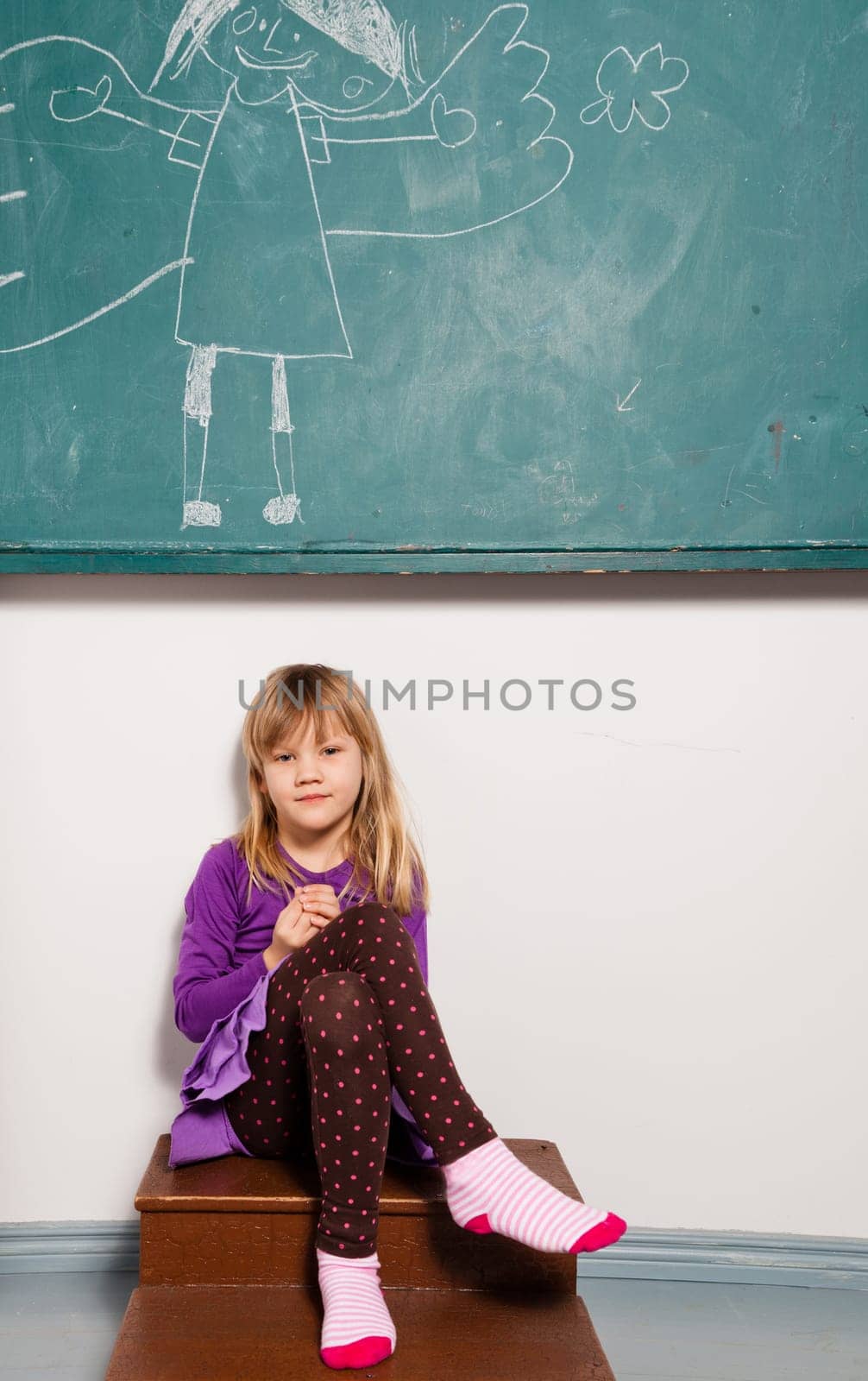 Studio portrait of relaxed young girl in classroom sitting in front of chalkboard with drawing of angel