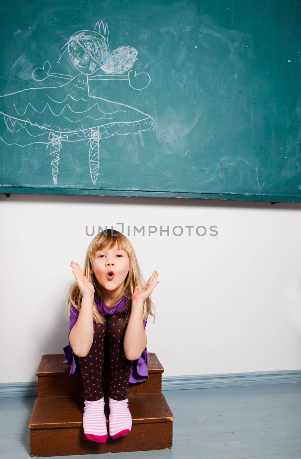 Studio portrait of young girl sitting in classroom in front of chalkboard with drawing and blowing kiss