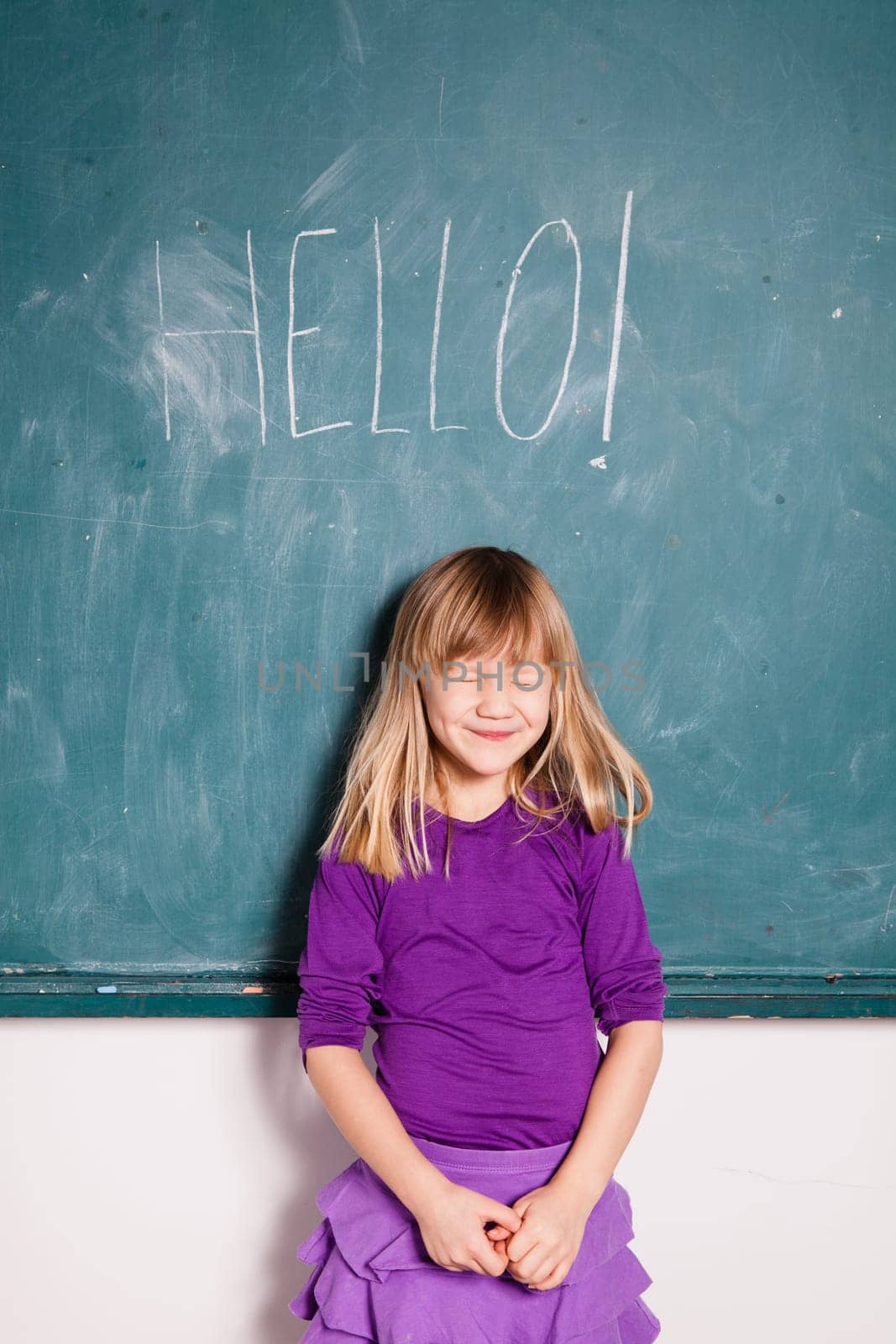 Studio portrait of smiling young girl with eyes closed in classroom with word hello written on chalkboard