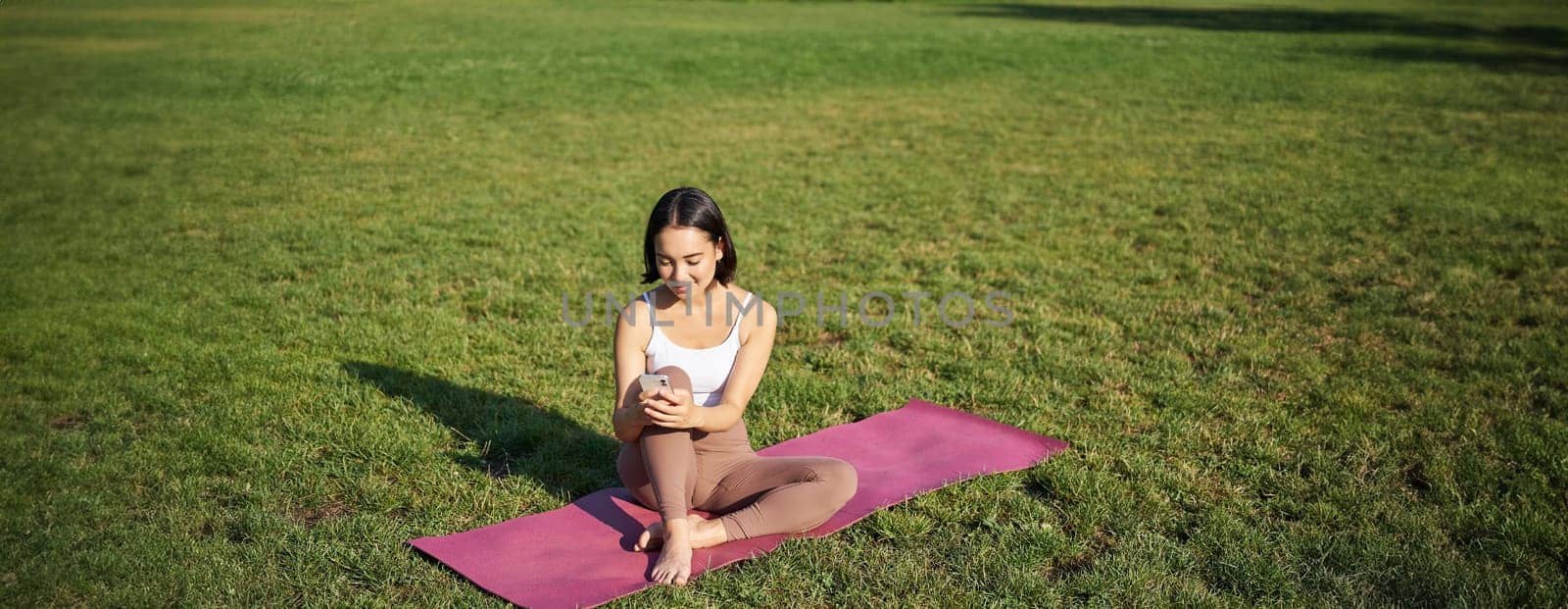 Woman in park, watching yoga video on smartphone, meditating on fresh air, sitting on rubber mat by Benzoix